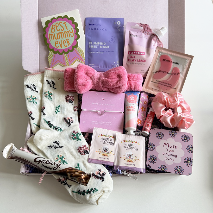 Open pink gift box arranged with mum pamper treats including cosy floral socks, skincare masks, fluffy pink spa headband, tea sachets, hand cream, chocolate bar, scrunchie and a “Best Mummy Ever” card.