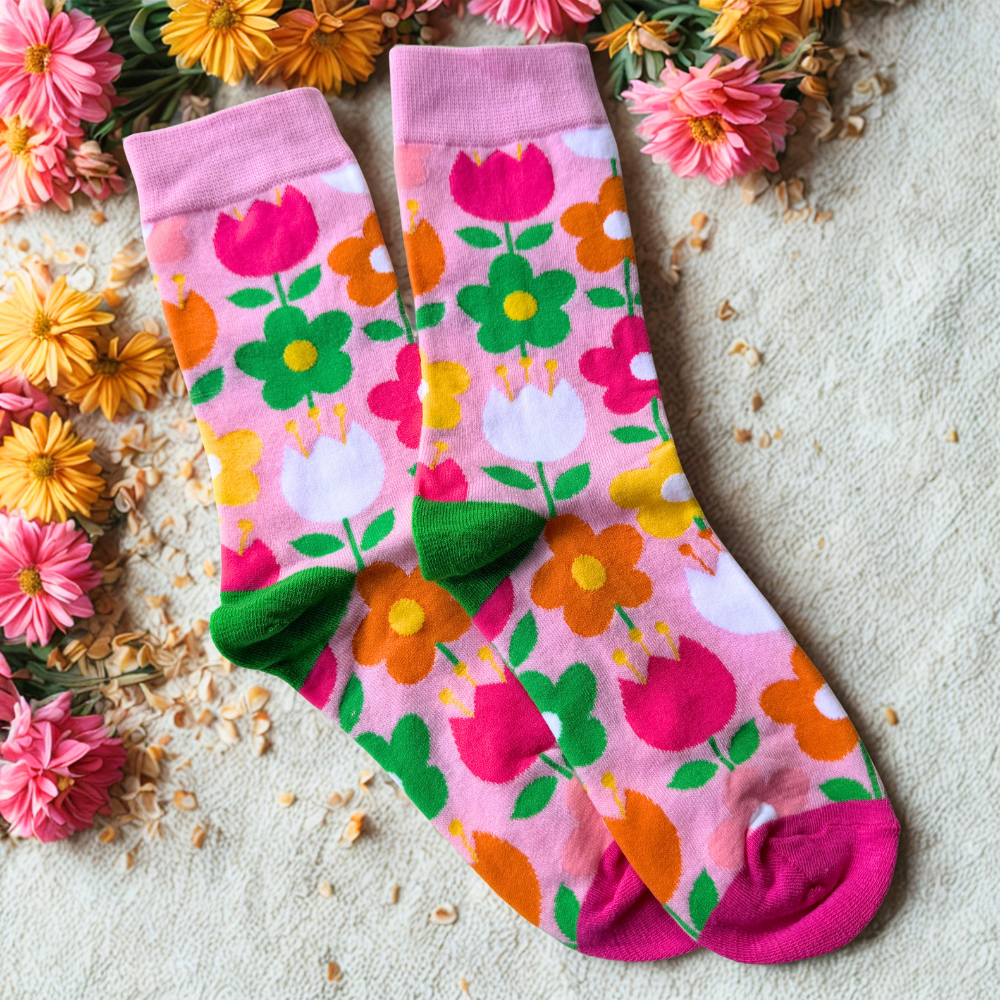 Pink socks featuring a colourful all-over floral pattern with bright pink, orange, white and green flowers. The socks have pink cuffs and toes with green heels and are displayed flat on a cream textured background surrounded by pink and orange flowers and scattered flower petals.