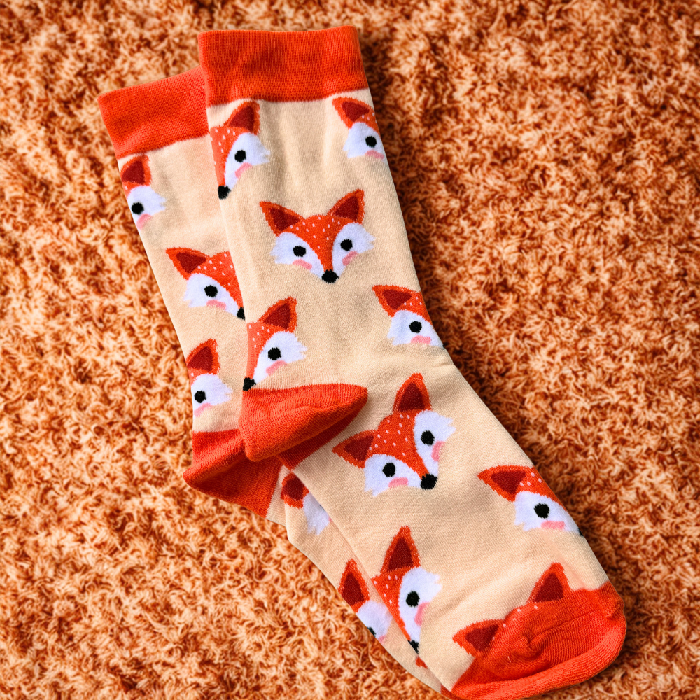 Cream and orange novelty socks featuring an all-over fox face pattern. The socks have bright orange cuffs, heels and toes, with cute orange fox faces scattered across a light cream background. The socks are displayed laid flat on a textured orange surface.