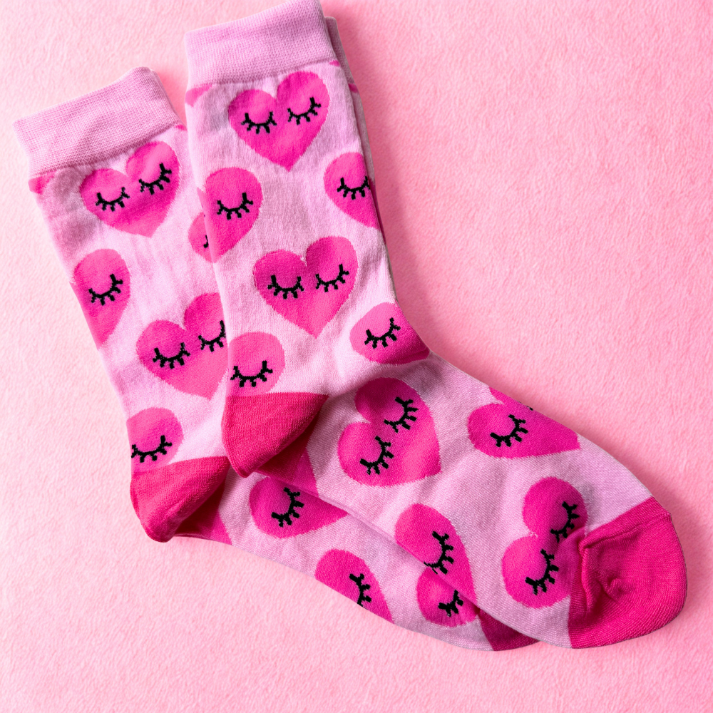 Light pink novelty socks featuring an all-over pattern of bright pink hearts with closed eyes and long eyelashes. The socks have matching bright pink cuffs, heels and toes and are displayed laid flat on a soft pink textured background.