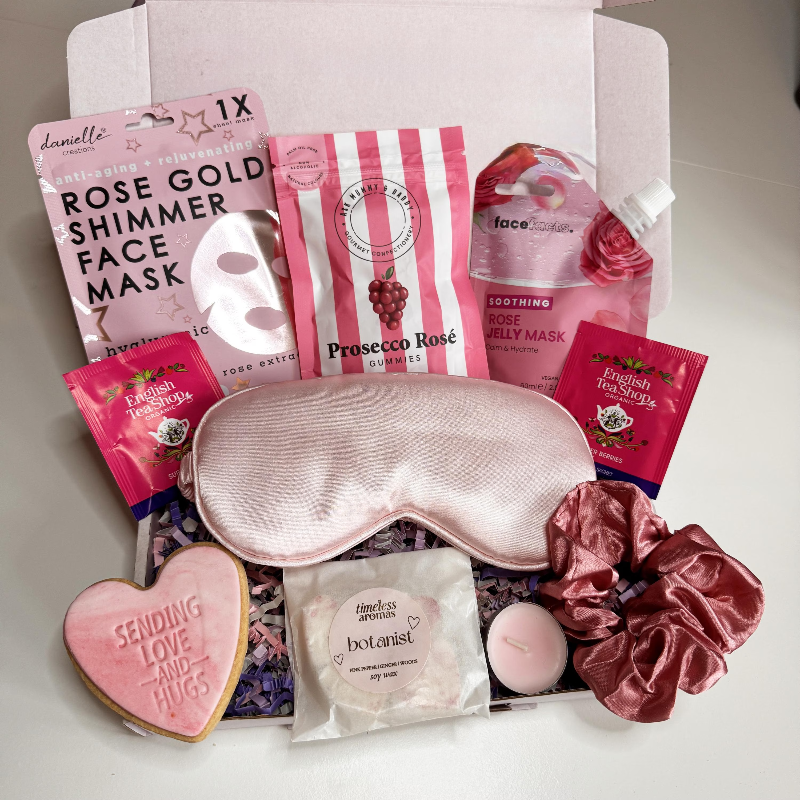 Open pink pamper gift box containing rose-themed beauty treats including sheet face masks, silk sleep mask, pink scrunchies, tea sachets, rose gummies, wax melt, tealight candle and a heart biscuit reading “Sending Love and Hugs.”