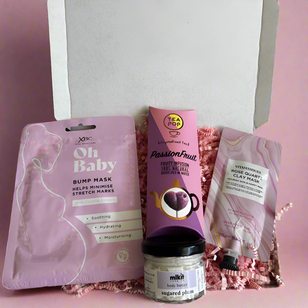 Pregnancy relaxation gift box with skincare and pampering items in pink packaging, including a clay mask, bump mask, body butter, and fruit tea, arranged in a gift box with shredded pink filler.