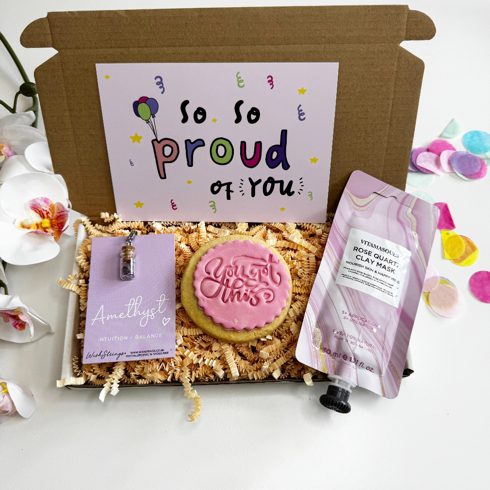 Motivational gift box with “So Proud of You” card, pink iced “You Got This” biscuit, rose quartz clay mask, and amethyst crystal pendant, displayed in pink shredded paper with colourful confetti.