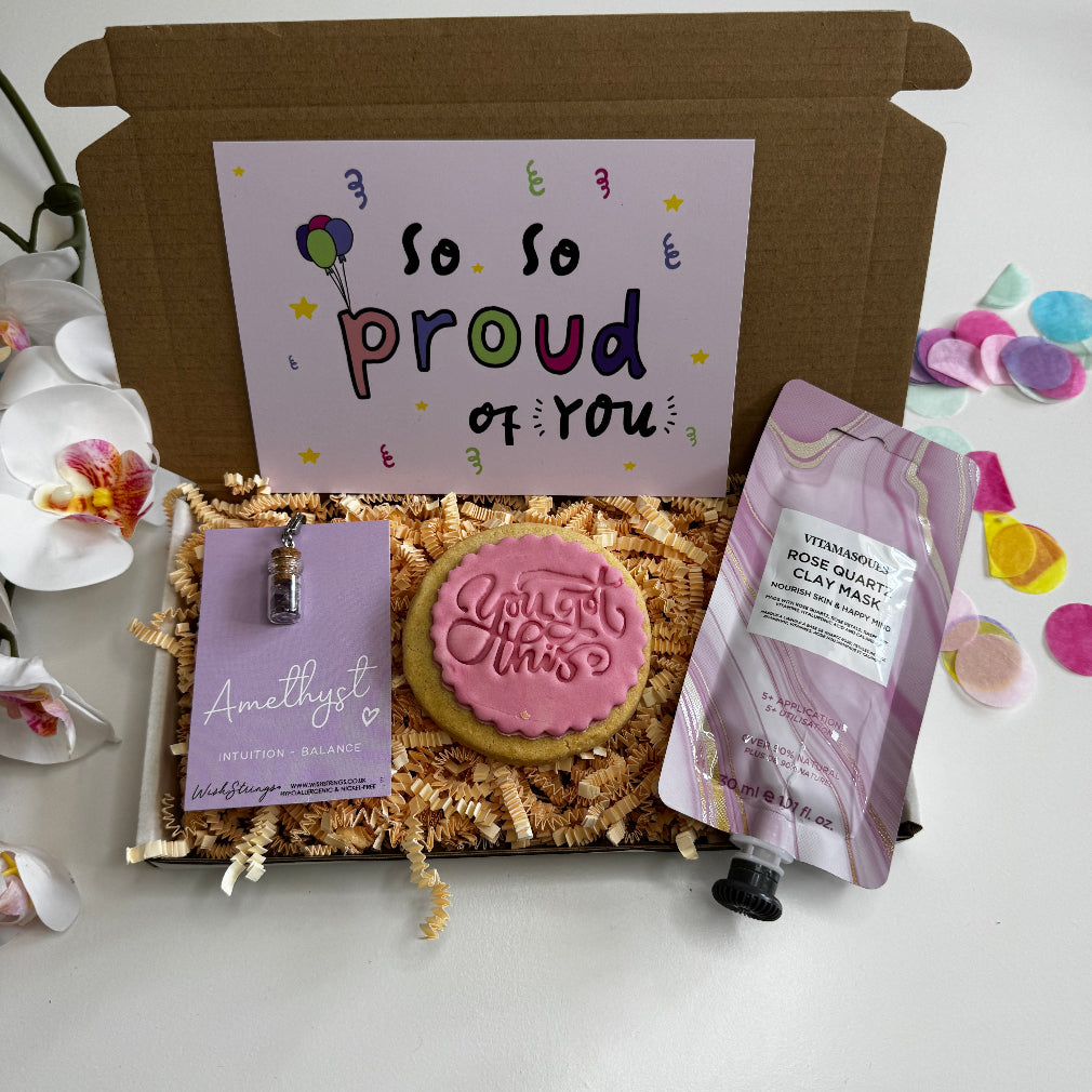 Motivational gift box with “So Proud of You” card, pink iced “You Got This” biscuit, rose quartz clay mask, and amethyst crystal pendant, displayed in pink shredded paper with colourful confetti.