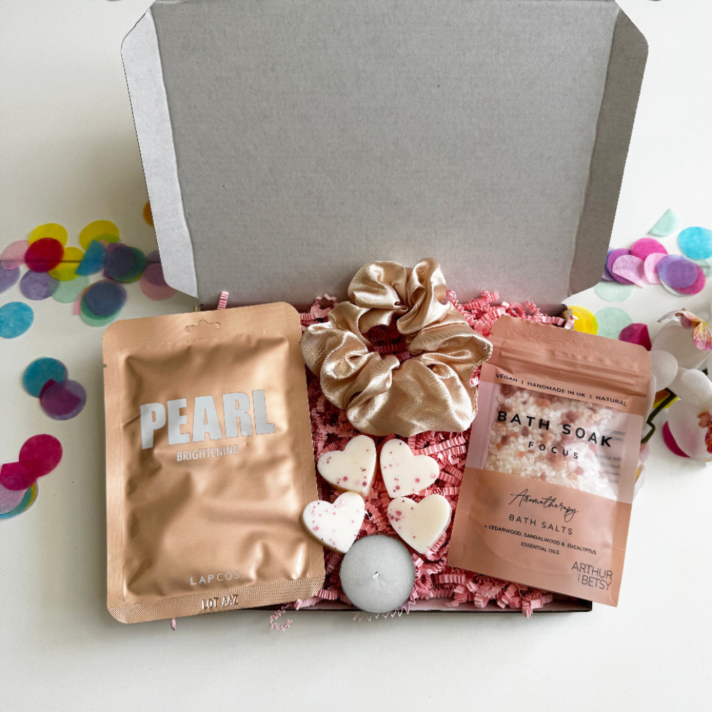 Relaxation gift box with pearl face mask, aromatherapy bath salts, ivory satin scrunchie, tealight candle, and heart-shaped wax melts, presented in pink shredded paper with confetti.